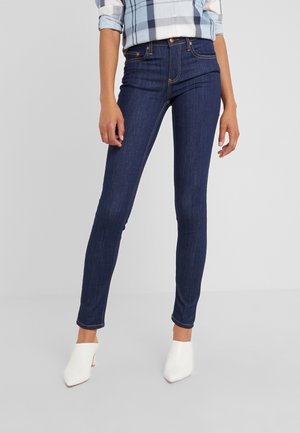 Person iført mørkeblå skinny jeans, en lyseblå ternet skjorte med opsmøgede ærmer og hvide højhælede sko står indendørs.