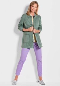 Groene suède jas met knoopsluiting, twee voorzakken; gecombineerd met lichtpaarse broek en zilveren loafers, tegen een witte achtergrond.