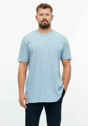 Mann mit Bart, der ein schlichtes hellblaues T-Shirt mit kurzen Ärmeln und eine dunkle Hose trägt, steht mit einer Hand in der Tasche vor weißem Hintergrund.