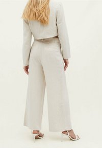 Blazer beige corto y pantalones de pierna ancha en tejido ligero. Corte notable en la espalda y dos bolsillos traseros en los pantalones. Sandalias de tacón alto blancas.
