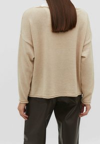 Beige gebreide trui met verlaagde schouders, geribde manchetten en een zoom. Draagt over donkerbruine synthetische broek, met een ontspannen pasvorm.