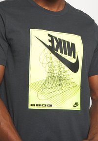 Mörkgrå bomull t-shirt med en stor neon-gul grafisk design och svart text "NIKE" i mitten, tillsammans med ett geometriskt mönster.