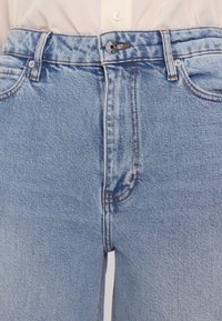 Jean en denim bleu clair taille haute avec fermeture par bouton et zip, porté avec une chemise beige rentrée.