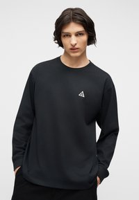 Νεαρό άτομο με σκούρα μαλλιά μέτριου μήκους φοράει μαύρο μακρυμάνικο πουκάμισο με μικρό λευκό τριγωνικό λογότυπο στο στήθος, με τα χέρια στις τσέπες.