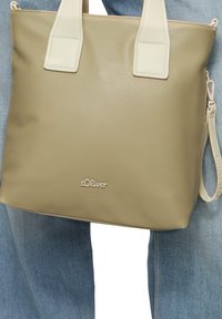 Beżowa torebka typu tote z ekoskóry z dwiema kremowymi uchwytami, z metalicznym logo z przodu oraz odpinanym paskiem.