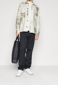 Camisa de manga larga de cuadros de colores claros con botones negros, llevada sobre una camiseta blanca; combinada con pantalones negros y zapatillas blancas, sosteniendo una bolsa de tela negra.