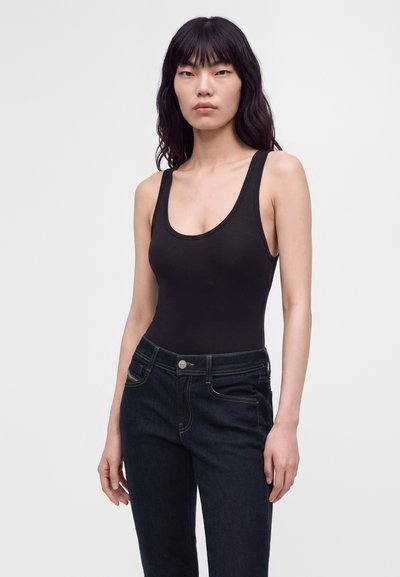 Femme aux longs cheveux noirs portant un haut noir sans manches et un jean bleu foncé, debout devant un fond clair uni.