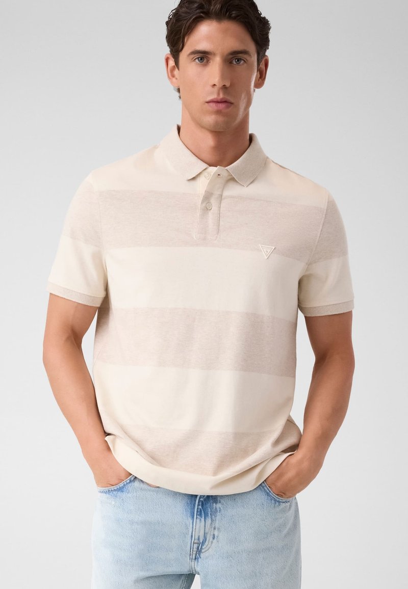 Homme portant un polo beige à rayures avec col et un jean bleu clair, debout les mains dans les poches devant un fond uni.