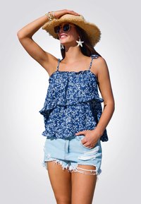 Mujer sonriendo, llevando un sombrero de paja, gafas de sol, una blusa sin mangas de flores azules, aretes de estrella de mar y shorts de denim claros rasgados, posando con una mano en el sombrero.