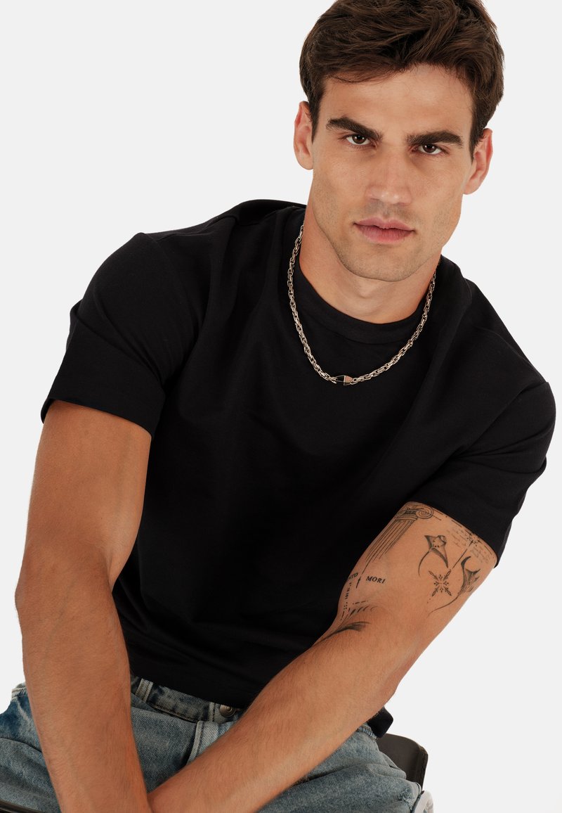 Sort bomulds t-shirt med korte ærmer, rund halsudskæring, båret med lyseblå denim jeans. Har et sølvfarvet kædehalssmykke og synlige tatoveringer.