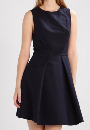 Kobieta ubrana w elegancką granatową sukienkę bez rękawów o długości do kolan, z dopasowaną talią i okrągłym dekoltem.