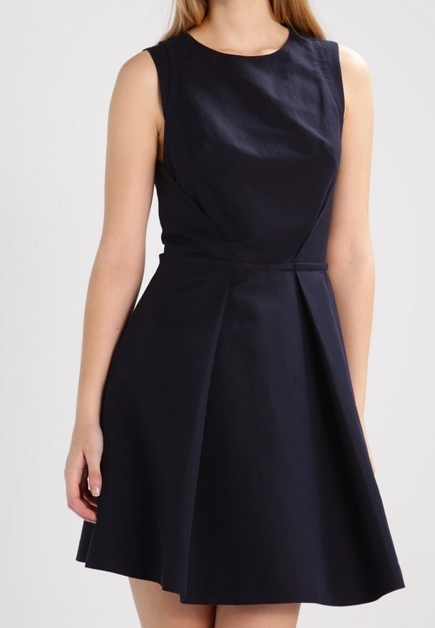 Femme portant une robe bleu marine sans manches, longueur au genou, coupe évasée en A avec une taille cintrée et un col rond.