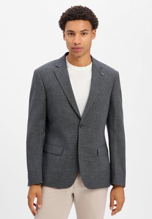 Jonge man met kort krullend haar, gekleed in een grijze blazer over een wit overhemd en beige broek, staand tegen een effen witte achtergrond.