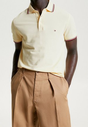 Polo en coton jaune clair avec col rayé rouge et bleu marine, patte de boutonnage à trois boutons, associé à un pantalon plissé beige.