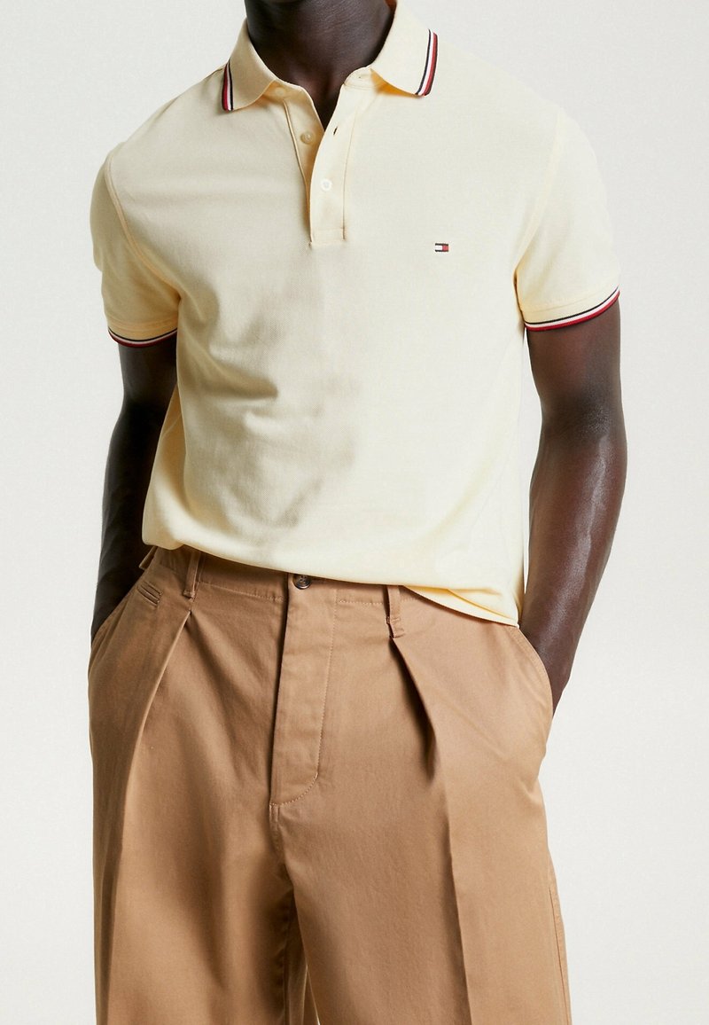 Polo en coton jaune clair avec col rayé rouge et bleu marine, patte de boutonnage à trois boutons, associé à un pantalon plissé beige.
