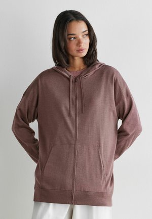 Giovane donna che indossa una felpa con zip mauve ampia e tasche frontali, che guarda di lato contro una parete liscia grigio chiaro.