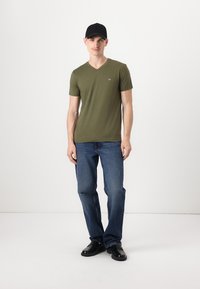 Olivno zeleno v-neck majica, slim fit, kratki rokavi; v kombinaciji s poštrihanimi modrimi kavbojkami in črnimi čevlji; nosi črno kapo. Preprost, sproščen videz.
