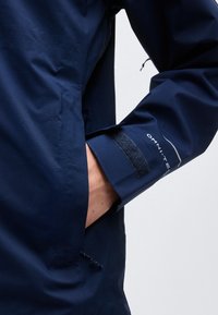 Άτομο που φοράει σακάκι σε μπλε ναυτικό χρώμα με το χέρι στην τσέπη του σακακιού, εμφανίζοντας την μανσέτα του μανικιού με ιμάντα βέλκρο και το λογότυπο "Omni-Tech".