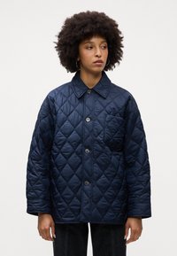 Giacca trapuntata blu navy con colletto strutturato, bottoni frontali e taschino sul petto con motivo a rombi obliqui.