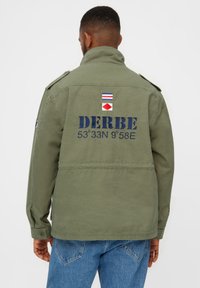Grøn militærstil jakke med høj krave. Har store marineblå bogstaver "DERBE" og geografiske koordinater på ryggen, sammen med broderede detaljer.