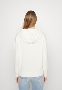 Valkoinen huppari pehmeällä pinnalla, jossa on kengurupocket, uritetut hihansuut ja helma. Yhdistetty vaaleansinisiin farkkuihin.