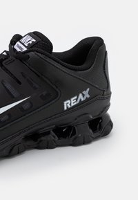 Zapatilla deportiva negra con un diseño elegante, materiales de malla y sintéticos, acentos texturizados y un logo prominente de "REAX".