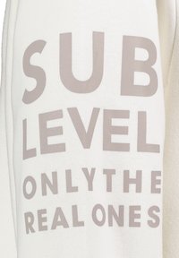 Weiße Sweatshirt mit großen grauen Buchstaben, die "SUB LEVEL ONLY THE REAL ONES" anzeigen. Das Material hat eine weiche Textur, typisch für Fleece.