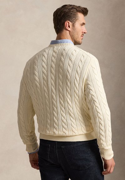 Uomo che indossa un maglione a trecce color crema sopra una camicia azzurra, mostrato di spalle su uno sfondo neutro.