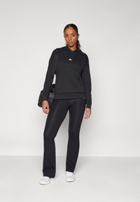 Moletão desportivo preto com bolsa canguru e pequeno logótipo, combinado com calças de treino justas pretas e ténis brancos, segurando uma mala no ombro.