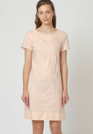 Joven mujer vestida con un vestido color melocotón claro con mangas cortas y bordados intrincados, posando frente a un fondo liso y claro.