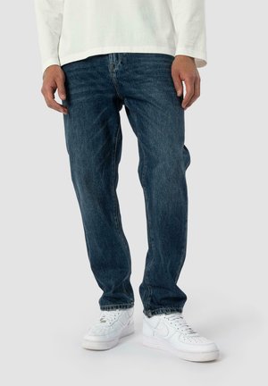 Dark blue denim jeans with a relaxed fit, featuring front pockets, subtle stitching, and worn texture. Paired with white sneakers.