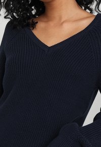 Pull en tricot bleu foncé avec col en V, texture côtelée et manches larges avec détails accentués aux poignets.