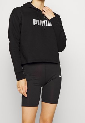 Personne portant un sweat à capuche noir Puma avec un logo métallique et un short de vélo noir Puma, debout devant un fond clair uni.