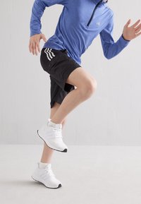 Persona che indossa una maglietta a maniche lunghe blu, pantaloni corti neri e sneakers bianche, colta in movimento con un ginocchio sollevato all'interno su un pavimento chiaro.