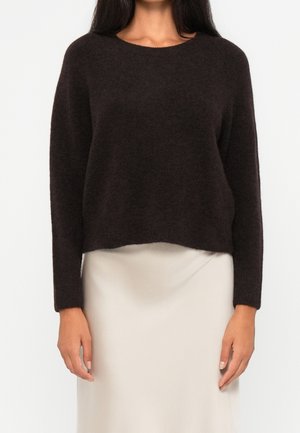 Femme portant un pull pelucheux marron foncé à manches longues associé à une jupe écrue, debout devant un fond clair uni.