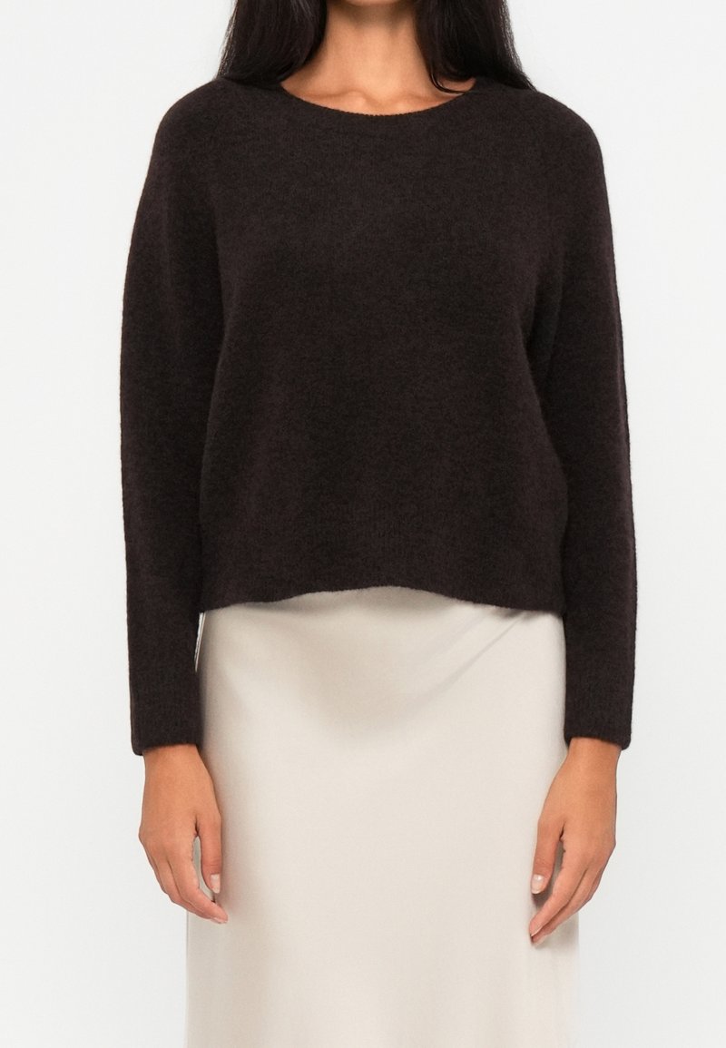 Femme portant un pull pelucheux marron foncé à manches longues associé à une jupe écrue, debout devant un fond clair uni.