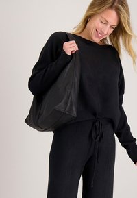 Zwarte oversized trui met geribbelde manchetten en een trekkoord in de taille, gecombineerd met bijpassende zwarte broek en een getextureerde zwarte shopper.