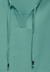 Haut à capuche bleu sarcelle clair avec une surface texturée, doté d'un col en V et de cordons avec des embouts noirs de chaque côté.