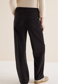Zwarte brede broek met achterzakken, subtiele stikdetails en een gladde textuur, gecombineerd met lichtgekleurde sneakers.