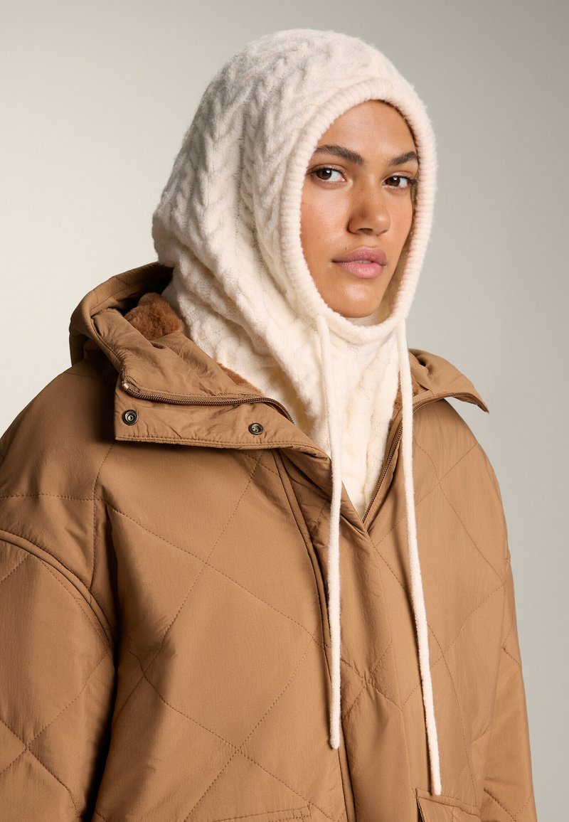 Bruine gewatteerde jas met oversized capuchon, gecombineerd met een crèmekleurige gebreide hoofddoek met textuurpatronen en trekkoorden.