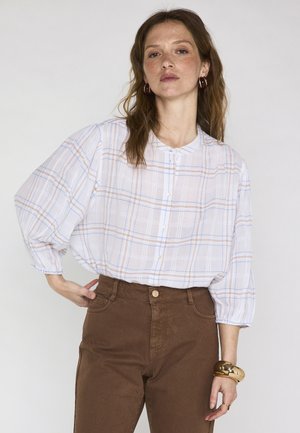 Donna che indossa una camicia bianca a quadri con bottoni e maniche a tre quarti, pantaloni marroni a vita alta, accessoriata con braccialetti dorati e orecchini a cerchio.