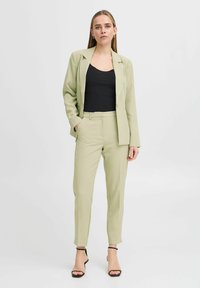 Svetlozeleni kostim z oblikovanim sakoom in zoženimi hlačami. Črna majica pod njim. Gladka tkanina, minimalno podrobnosti in petke s gležnjevimi trakovi.