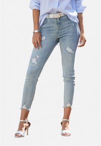 Jeans skinny azzurri chiari con effetto vissuto, orlo sfrangiato e multiple strappi. Abbinati a tacchi bianchi open-toe e una camicia a righe chiare.
