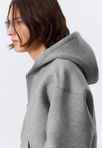 Harmaa vetoketjullinen huppari, valmistettu pehmeästä kankaasta, jossa on suuri huppu ja rento istuvuus. Näkyvät saumat ja kokonaan vetoketjulla varustettu etuosan yksityiskohta.