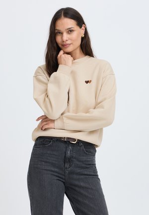 Jeune femme aux longs cheveux foncés portant un sweat beige avec deux petits motifs en forme de cœur et un jean noir, posant avec la main près du menton.