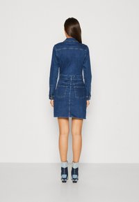Dżinsowa sukienka z kołnierzykiem, długimi rękawami i dopasowaną talią. Ciemnoniebieski kolor, dwie tylne kieszenie i dół kończący się w połowie uda.