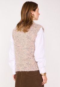 Pull sans manches rose pâle en tricot, présentant une texture tachetée et un ourlet côtelé, porté par-dessus une chemise blanche à manches longues.