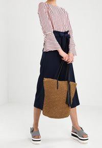 Bluzka w paski z długim rękawem w czerwono-białe pasy, granatowe spodnie z wysokim stanem, pleciona brązowa torba na ramię z frędzlami oraz czarno-białe sandały na platformie w paski.