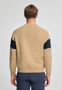 Felpa beige con collo a giro, maniche raglan e dettagli neri sulle maniche superiori. Il tessuto è morbido e ha una texture liscia.