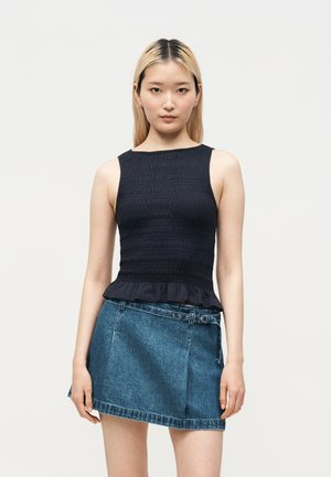 Donna con capelli biondi lisci che indossa un top nero senza maniche e una minigonna di jeans blu con cintura, su sfondo bianco.